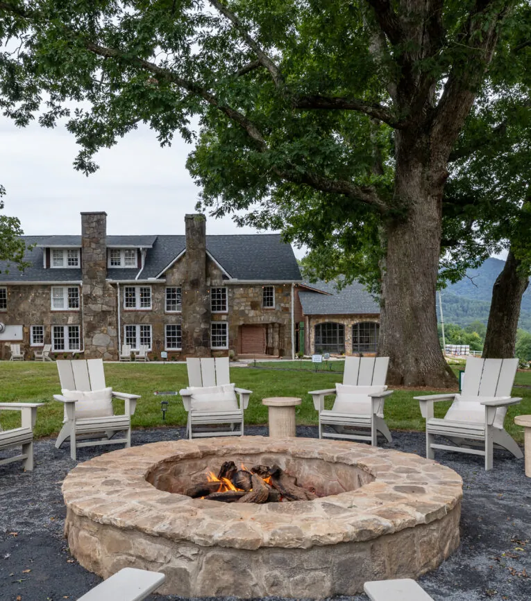 Stone fire pit with burning fire, surrounded by white chairs, sits under large trees in front of a stone house and lush green lawn.