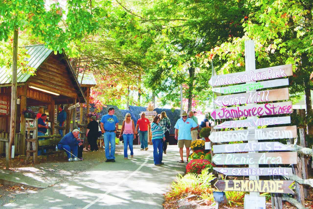 People walk along a tree-lined path at a festival, with colorful wooden signs pointing to various attractions like crafts, lemonade, and a museum.