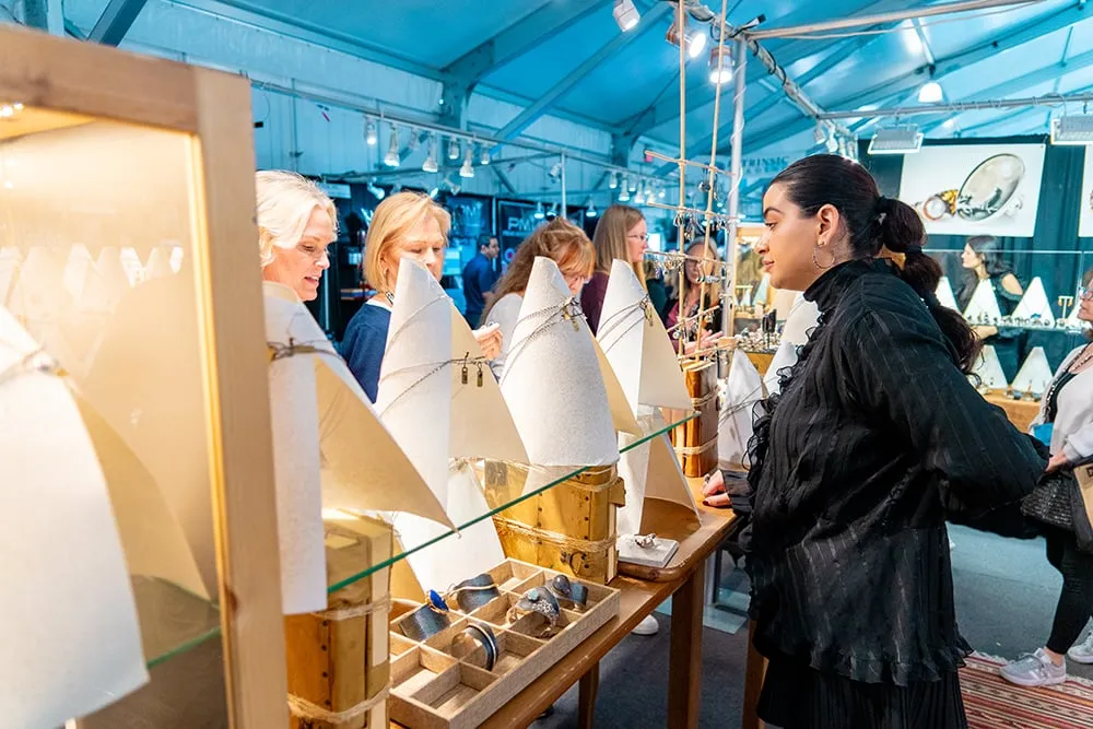 Women browse and discuss jewelry displayed on stands at an indoor market or craft fair under a large, well-lit tent.