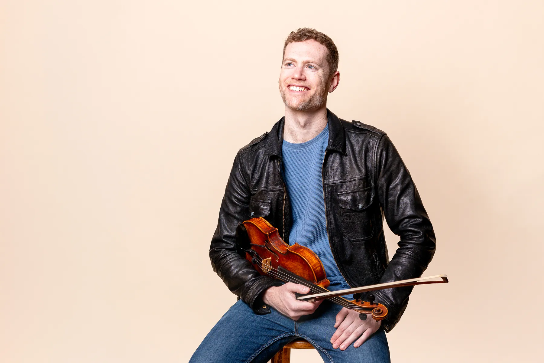 A smiling man in a black leather jacket and blue shirt sits on a stool, holding a violin and bow against a light beige background.
