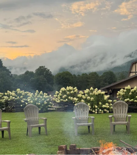 Four empty lawn chairs by a fire pit overlook misty mountains and a barn at sunset—near hotels in downtown Dillard House, PA.