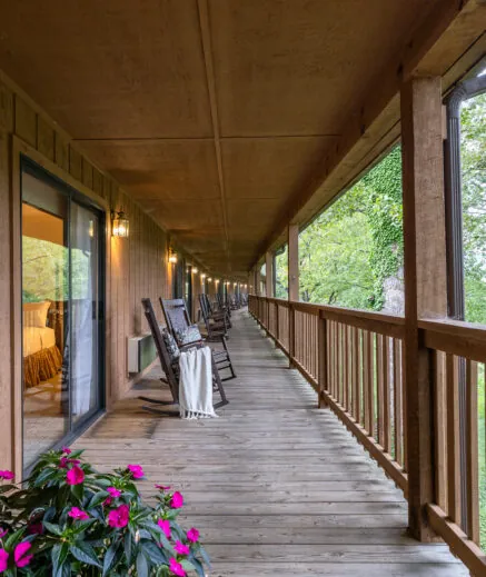 A long wooden porch with rocking chairs, a railing, and potted pink flowers, overlooking green trees and a warmly lit room through glass doors.
