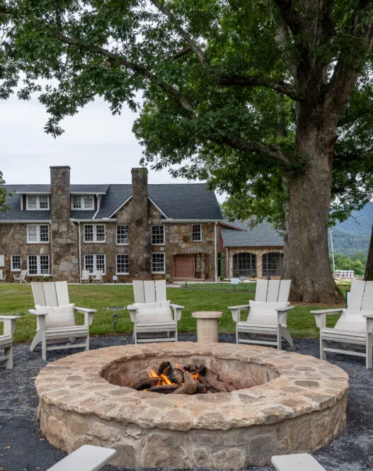 White Adirondack chairs surround a fire pit near a stone house, with mountain views—perfect for guests at hotels in downtown Dillard House PA.