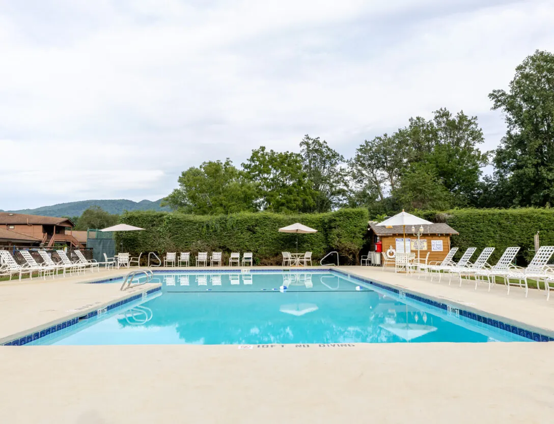 Outdoor swimming pool with clear water, surrounded by white lounge chairs, umbrellas, and trees under a cloudy sky.