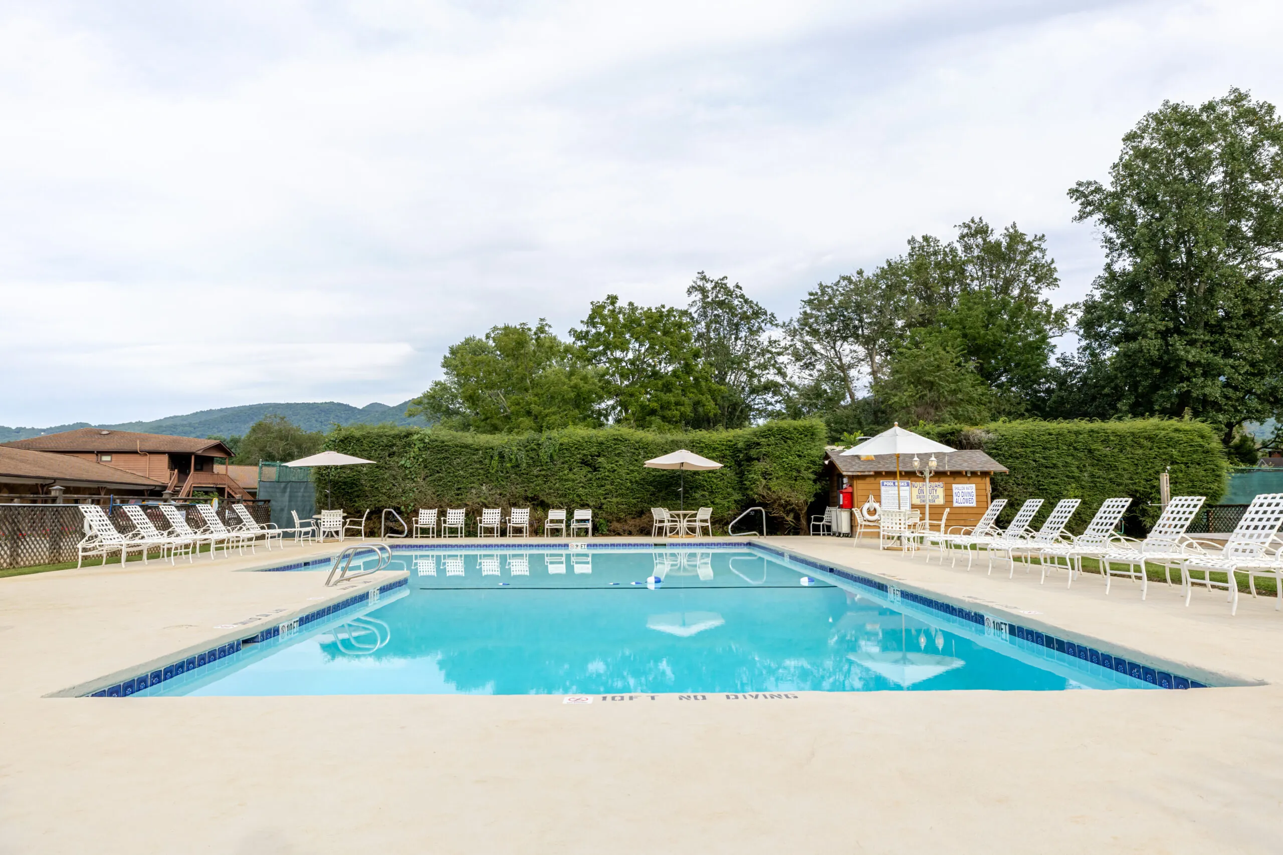 Outdoor swimming pool with clear water, surrounded by white lounge chairs, umbrellas, and trees under a cloudy sky.