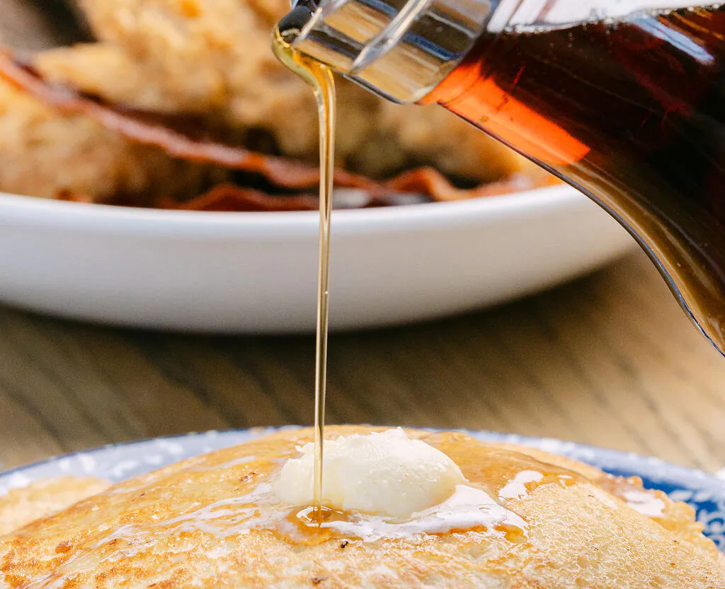 Syrup being poured from a glass pitcher onto a stack of pancakes topped with a pat of butter, with a plate of bacon in the background.