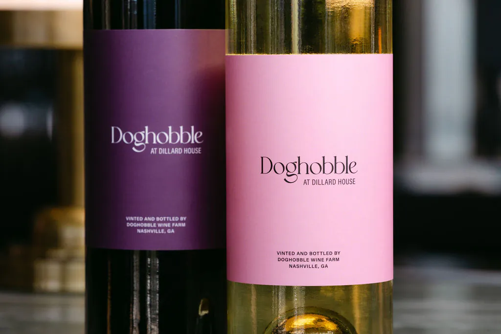 Two wine bottles labeled Doghobble at Dillard House, one with a purple label and one with a pink label, standing side by side on a table.