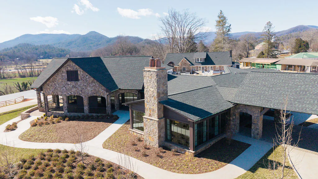 Aerial view of a large stone and wood building with mountain scenery in the background and landscaped walkways in front.