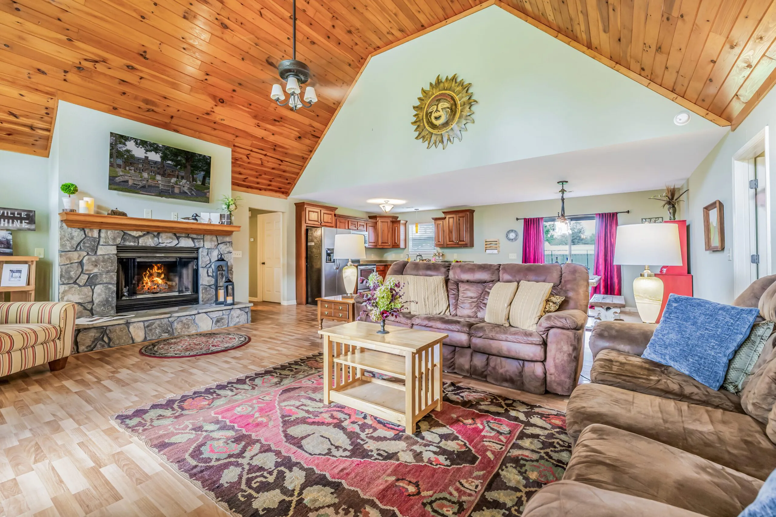 Cozy living room with stone fireplace, brown sofas, wood ceiling, colorful rug, and open kitchen in the background with wooden cabinets.