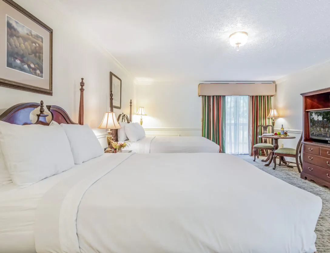 Bright hotel room with two large beds, wooden furniture, a TV, lamps, a window with curtains, and a small table with chairs.