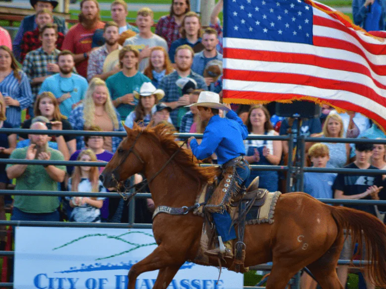 A rodeo rider in a blue shirt and white hat competes on a brown horse in front of a crowd and an American flag.