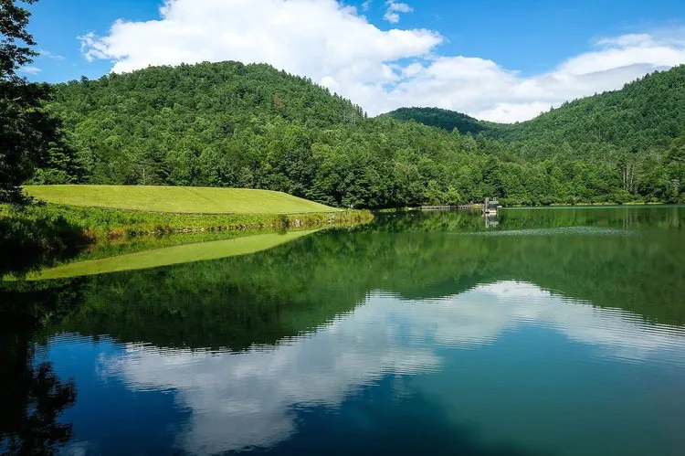A calm lake reflects green hills, trees, and a partly cloudy blue sky on a bright day.