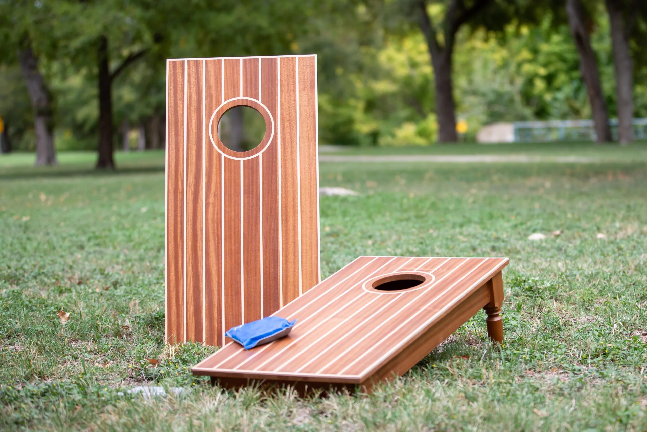 Two wooden cornhole boards on grass in a park, with one blue bean bag resting on the front board. Trees are visible in the background.