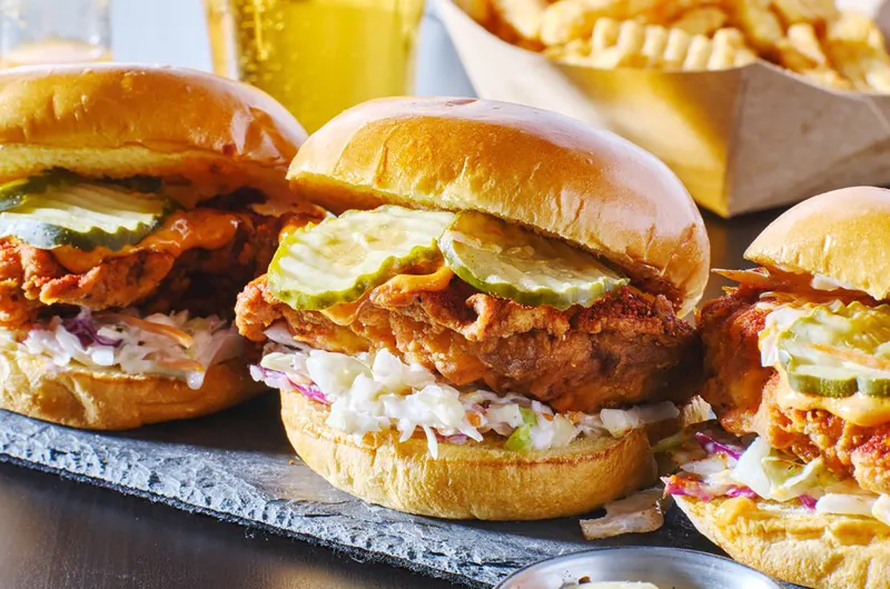 Three fried chicken sandwiches with pickles and coleslaw on buns, served on a slate, with fries and a drink in the background.