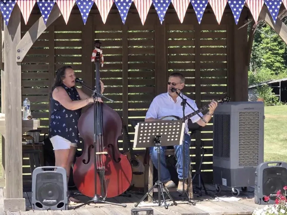 A woman plays an upright bass and sings with a man on guitar under a gazebo decorated with American flag bunting.