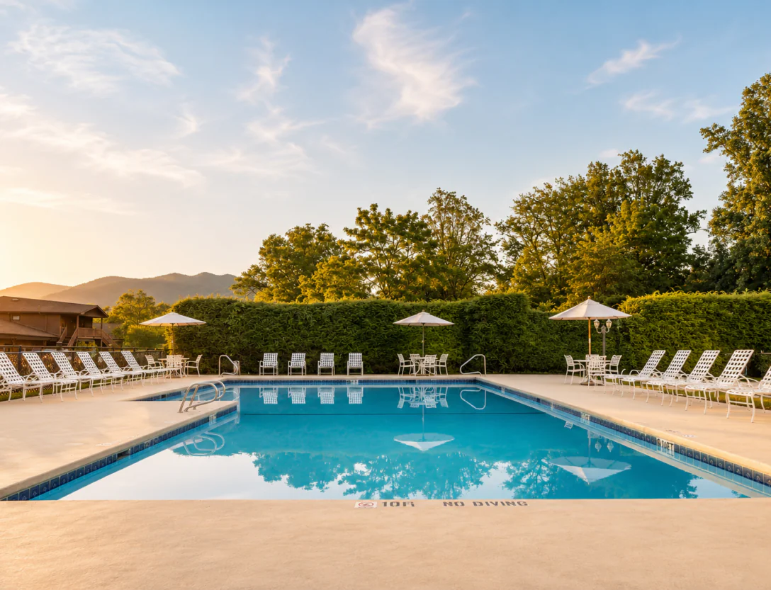 Outdoor swimming pool surrounded by white lounge chairs and umbrellas, with trees and hills in the background at sunset.