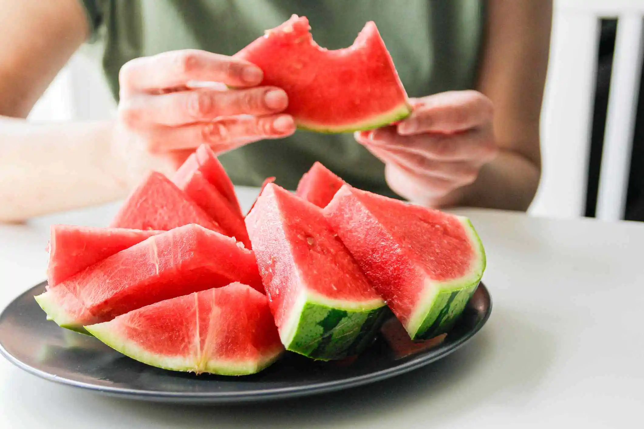 A person holds a slice of watermelon with a bite taken out, while several more watermelon slices sit on a plate in front of them.