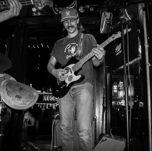 Man wearing a cap and t-shirt plays electric guitar on stage at a live music venue, with lights and signs in the background. Black and white photo.