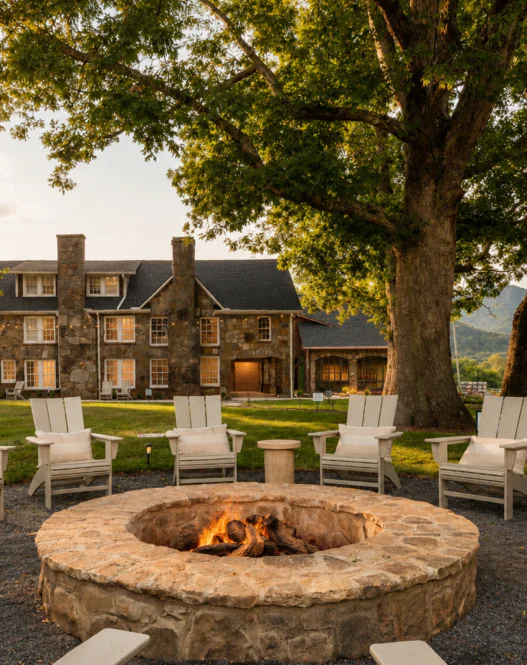 A stone fire pit with Adirondack chairs sits under large trees, facing a rustic house and green lawn, with mountains in the background at sunset.