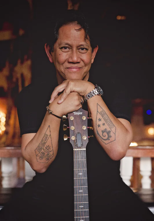A man with tattoos on both forearms rests his hands on the headstock of a Taylor guitar.